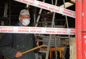 A man works on a construction site. White tape saying "WARNING ASBESTOS REMOVAL KEEP OUT" can be seen. The man is wearing a protective mask.