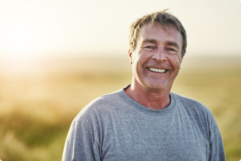 An older man wearing a grey shirt smiles in a field.
