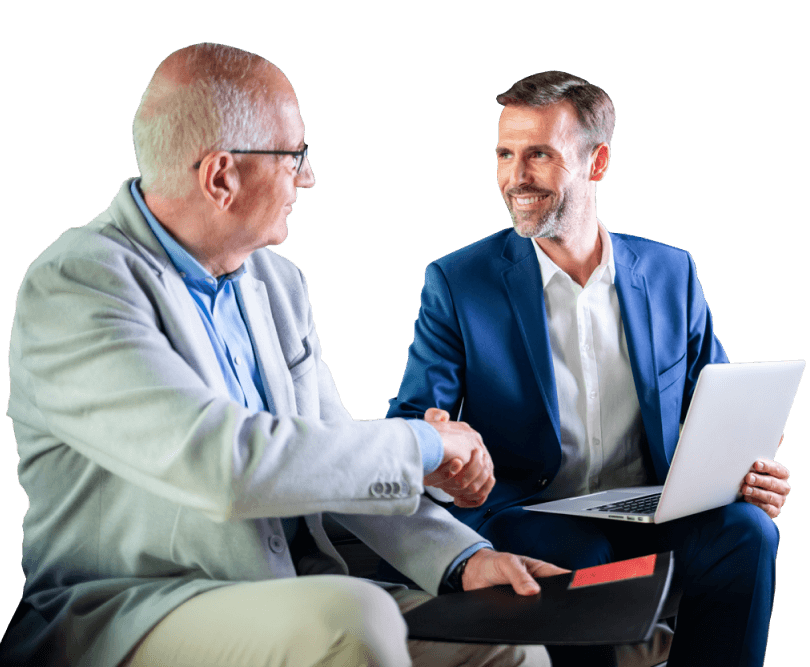 An older male shakes hands with a younger, professionally dressed male with a laptop.