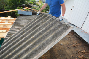 A worker holds a sheet of grey asbestos outside.