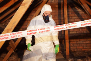 A worker in a hazmat suit works in a wooden building. Caution tape reading "Warning asbestos removal. Keep out" is seen in the foreground
