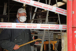 An older man removes asbestos from a building. Caution tape is draped in front of the building's entrance.