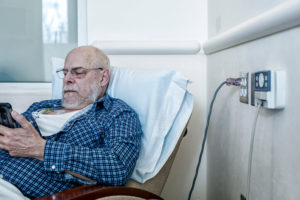 An older person sits in a hospital and stares at his phone.