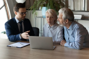 A younger man in a suit sits with an older couple at a desk. All three are looking at a grey laptop that's sitting on the desk.