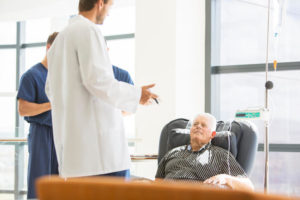 A doctor looks at an older male patient who is sitting in a chair getting chemotherapy
