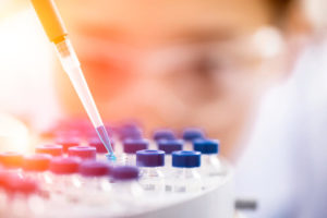 Closeup of a beaker dropping a blue liquid into a vial with a scientist in the background