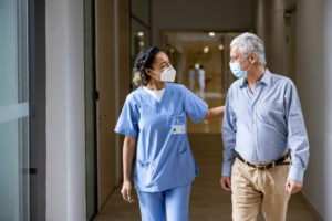 A female nurse in scrubs walks and pats the back of an older male patient. Both are wearing facemasks.