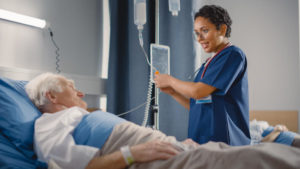 A nurse tends to an IV bag of an older male hospital patient who is sitting in a bed