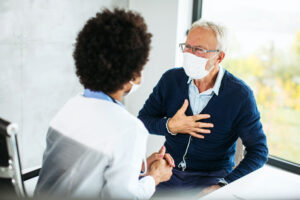 An older male patient in a face mask speaks with a younger person in a medical coat