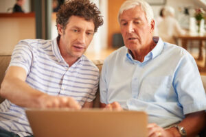 A young man and old man sit on a couch and look at a computer screen together