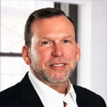 Ricky LeBlanc wears a black suit and white shirt in front of a white wall and window.